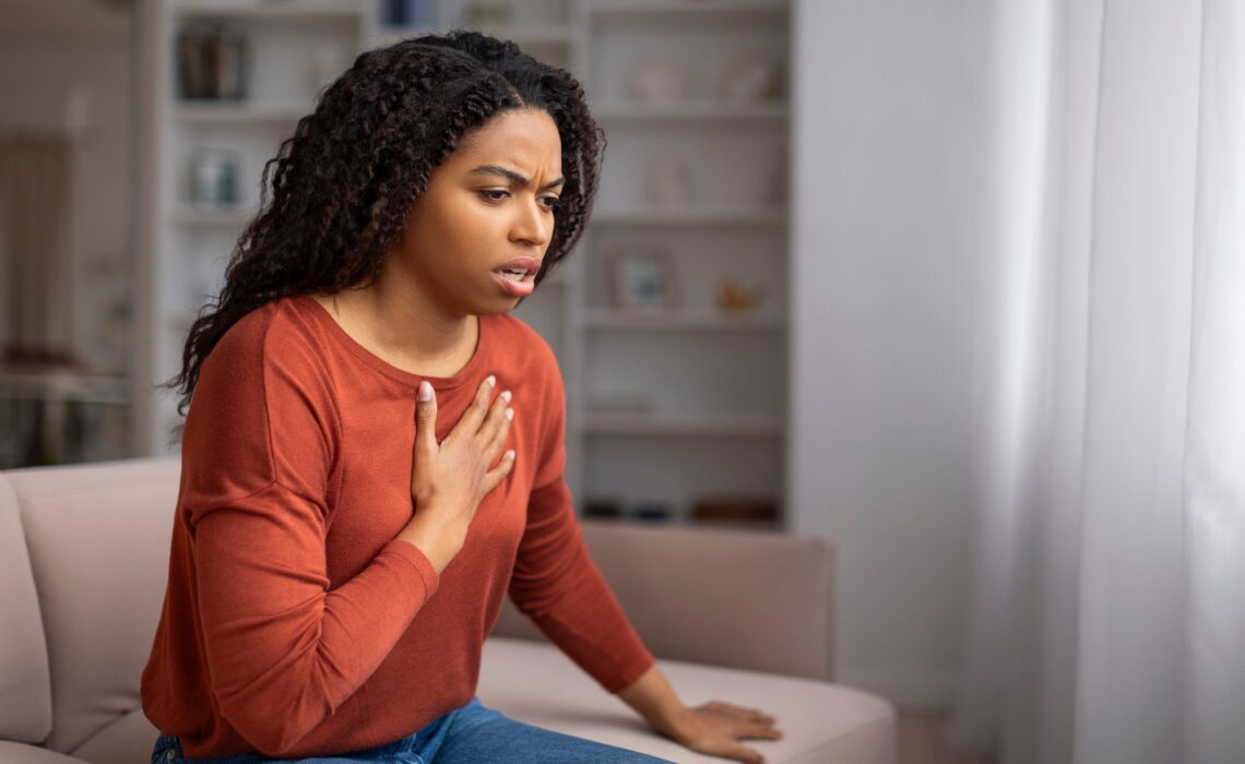 Concerned young black woman suffering pain in chest while sitting on couch